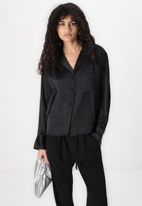 Zwarte satijnen blouse met lange mouwen en knopen aan de voorkant, een enkele borstzak en een zachte glans, gecombineerd met een zwarte broek en een zilveren clutch.
