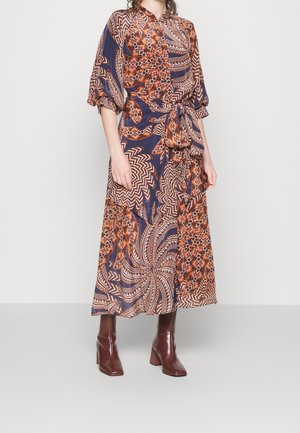 Kvinde iført en lang mønstret kjole med geometriske og swirl-designs i orange, marineblå og beige, kombineret med brune læder ankelstøvler.