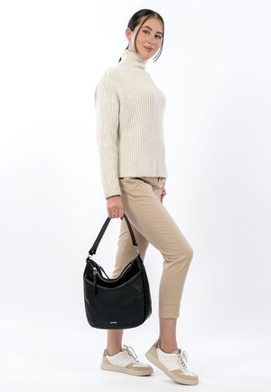 Kobieta w beżowych spodniach, kremowym dzianinowym golfie i trampkach, trzymająca czarną skórzaną torebkę na ramię, stojąca na białym tle.