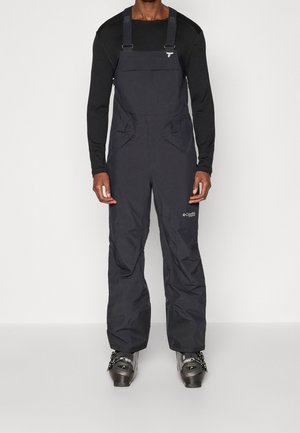Mann trägt ein schwarzes Langarmshirt, schwarze Columbia wasserdichte Latzhose und graue Skistiefel, steht vor einem weißen Hintergrund.