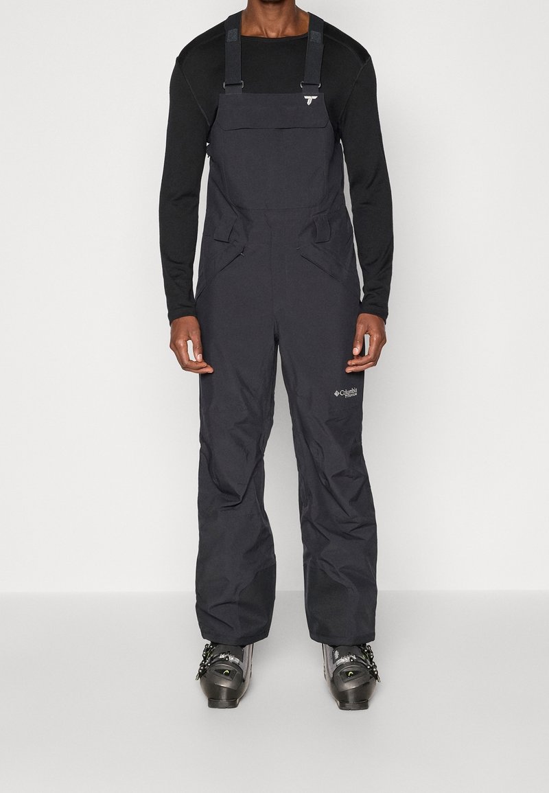 Homme portant une chemise à manches longues noire, un pantalon salopette étanche Columbia noir et des chaussures de ski grises, debout devant un fond blanc.