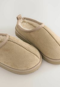 Zapatillas de ante beige con un forro suave y esponjoso, detalles de costura y una pestaña de bucle en la parte trasera para ponerlas y quitarlas fácilmente. Suela de goma plana.
