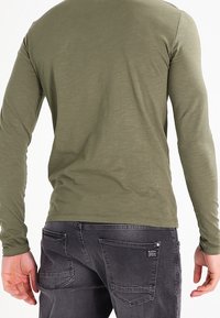 Camisa de manga larga de color verde oliva con textura suave y cuello redondo, combinada con vaqueros de color gris oscuro que tienen bolsillos traseros y un pequeño detalle de logo.