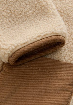 Tessuto in fleece di colore crema con superficie testurizzata, dotato di un polsino in corduroy marrone; finitura liscia e texture strettamente intrecciata nella cucitura.