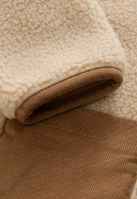 Fleece pulóver s textúrovaným krémovým telom a kontrastným hnedým manžetom z kordovej látky. Mäkký materiál s útulným, teplým vzhľadom.
