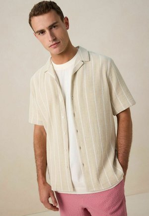 Short-sleeve beige shirt with vertical white stripes, textured fabric, and a collar, worn over a white T-shirt and paired with pink shorts.