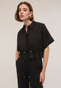 Blusa nera cropped in tessuto leggero, con maniche corte, tasca frontale, vita regolabile con cordoncino e dettagli metallici.