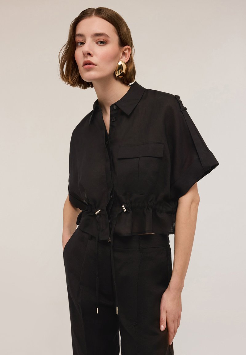 Blusa nera cropped in tessuto leggero, con maniche corte, tasca frontale, vita regolabile con cordoncino e dettagli metallici.