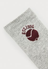 Grau gestrickte Socke mit einem gerippten oberen Abschluss, die ein burgunderfarbenes Rundlogo zeigt, das "EST 1954" und ein Tennisracket-Motiv beinhaltet. Weicher Strickstoff.