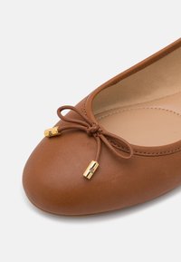 Hnědé kožené baletní bačkory s zaoblenou špičkou, zdobené mašlí a zlatými kovovými doplňky na tkaničkách. Hladký povrch s jemným leskem.