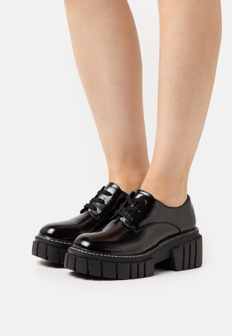 Chaussures à plateforme épaisses noires brillantes à lacets, portées sur des jambes nues, photographiées sur un fond blanc.