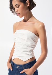 Weißes trägerloses Oberteil mit geraffter Textur, figurbetontem Design und spitzem Saum, kombiniert mit einer dunklen Jeanshose mit Knopfverschluss.