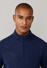 Granatowy sweter na druty z wysokim kołnierzem i ćwierć zamkiem błyskawicznym. Posiada fakturowany wzór w warkocze oraz subtelny detal z logo na piersi.