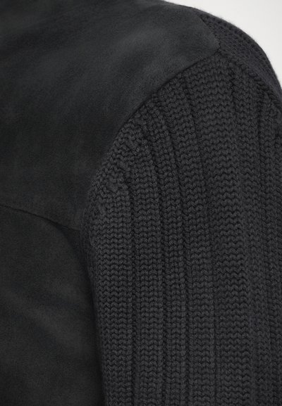 Sweater noir avec une combinaison de daim lisse et de texture tricotée côtelée sur les manches, mettant en avant des matériaux contrastants.
