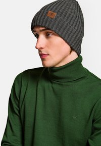 Szary rybaczasty beanie z skórzaną łatką, noszony z zielonym swetrem z wysokim kołnierzem. Beanie ma dopasowany krój i teksturowany splot.