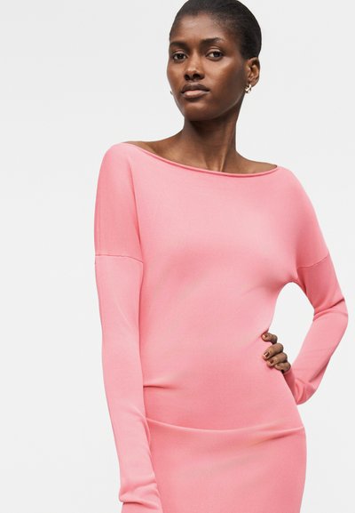 Μια γυναίκα φοράει φόρεμα σε ροζ χρώμα με μακριά μανίκια και ακάλυπτους ώμους, ενώ έχει το χέρι της ακουμπισμένο στο ισχίο, σε ένα απλό φόντο.