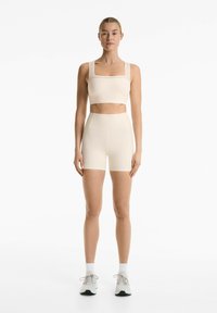 Beige sports bra with square neckline and decorative detailing, paired with high-waisted beige shorts. Fabric appears smooth and stretchy.