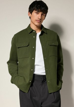Dark green jacket with a button-up front, two chest pockets, and a collared neckline, paired with a white shirt and black trousers.