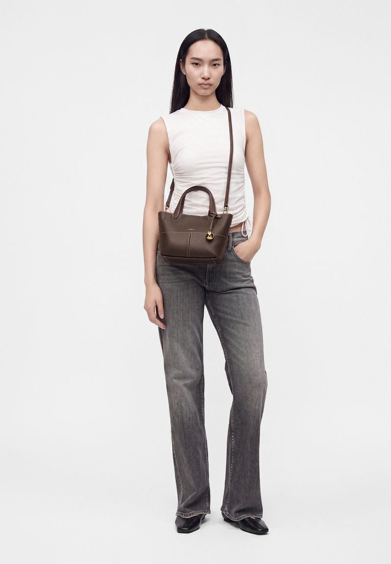 Jeune femme portant un haut blanc sans manches, jean gris, chaussures noires, portant un petit sac à main marron avec une bandoulière, debout devant un fond blanc.