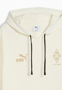 Hanorac de culoare crem, cu snururi negre, un logo pe piept și accente cu textul "KING" și "125 JAHRE" în aur. Material moale.