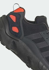 Black athletic shoe featuring a textured rubber sole, mesh upper, and red accents on the heel and side. Reinforced sections for durability.