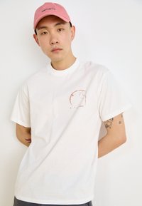 White cotton t-shirt with short sleeves, featuring a reddish abstract design on the left chest area. Smooth texture, relaxed fit.