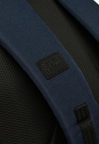 Granatowy pasek plecaka z teksturowanej tkaniny i siateczkowym podszyciem, z czarną kwadratową łatą z logo oznaczoną "SUPERDRY".