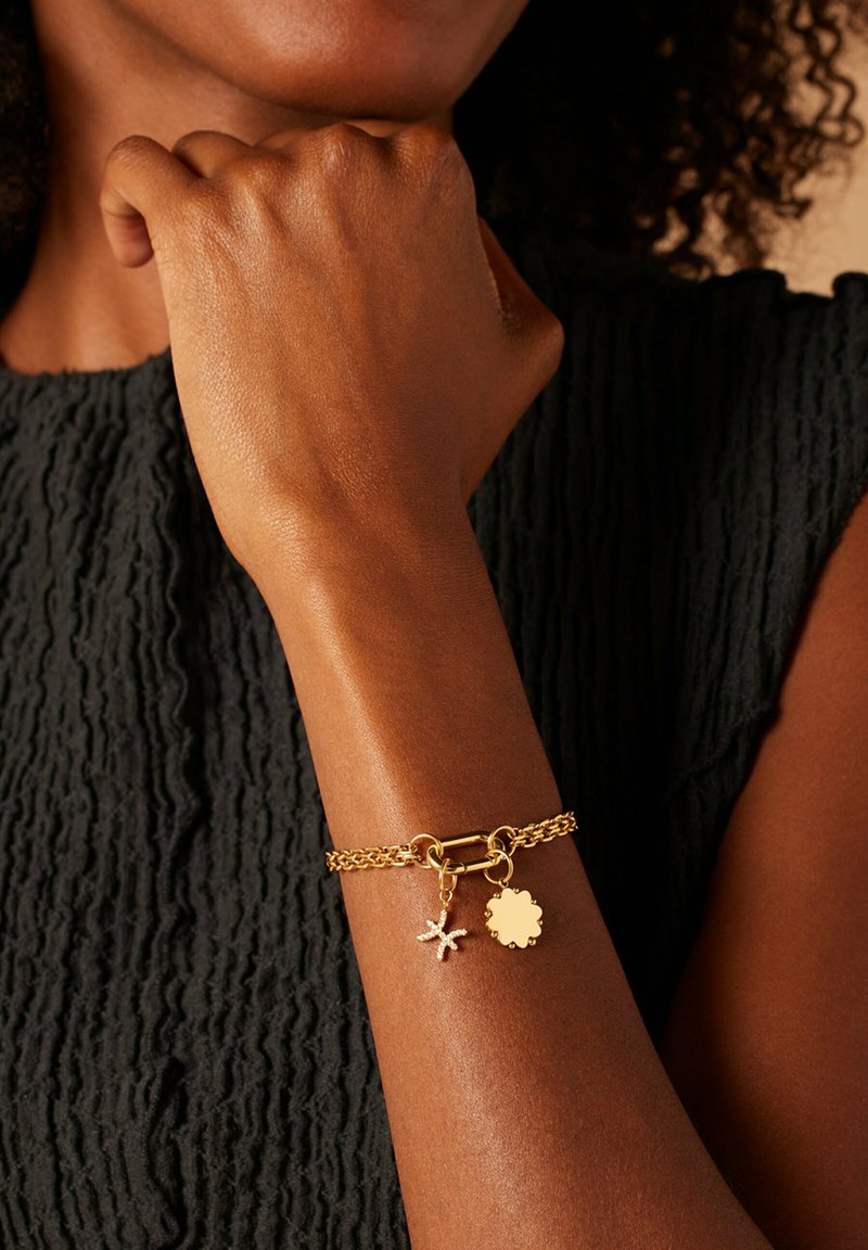 Gouden kettingarmband met zeester- en ronde bedeltjes, voorzien van een textuurdetail en een veilige sluiting. Gelegen op een gladde huidkleur.