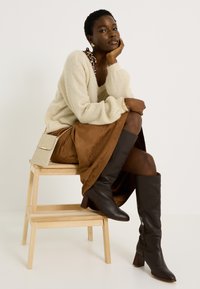 Mujer con el cabello corto, vestida con un suéter crema, falda marrón, botas altas marrón oscuro y un pequeño bolso brillante, sentada en taburetes de madera.