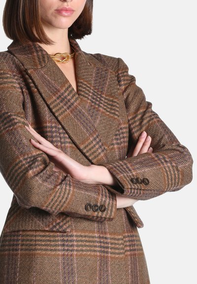 Femme portant un blazer à carreaux marron et un collier chaîne torsadée en or, bras croisés visibles des épaules à la taille.