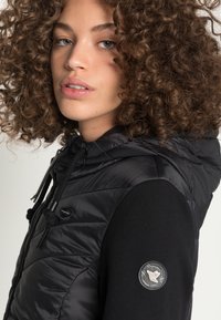 Gilet imbottito nero con cappuccio, dotato di chiusura con zip e cordino. Accentuato da una toppa con logo sulla manica nera.