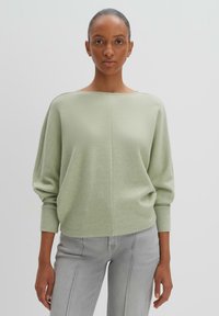 Pullover verde chiaro con scollatura ampia, spalle cadenti e maniche lunghe. Texture morbida e vestibilità ampia, abbinato a jeans grigi chiari.
