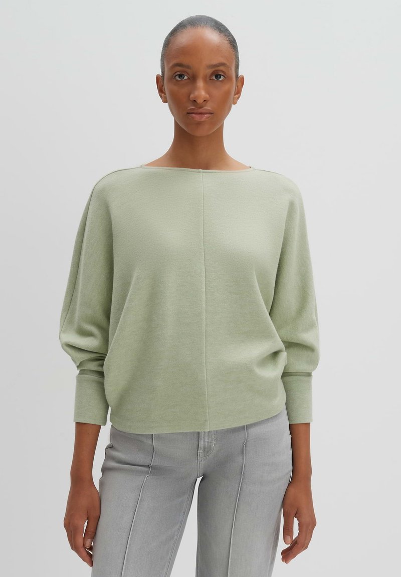 Pullover verde chiaro con scollatura ampia, spalle cadenti e maniche lunghe. Texture morbida e vestibilità ampia, abbinato a jeans grigi chiari.