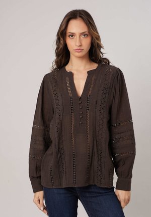 Vrouw draagt een donkerbruine blouse met lange mouwen, decoratieve uitsnijdingen en knopen, gecombineerd met blauwe jeans, staand tegen een effen achtergrond.