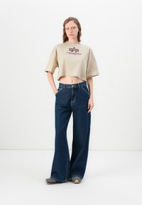 Beige kort t-skjorte med "Alpha Industries" logo, sammen med vide blå denim bukser. Skoene er grå med svarte detaljer.