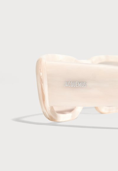 Слънчеви очила Jacquemus в кремав цвят с дебели квадратни рамки и лого на рамото на светъл фон.
