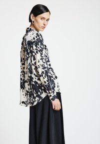 Blusa a maniche lunghe con fondo crema e macchie astratte nere, leggermente lucida, dalla vestibilità morbida e orlo arrotondato. Abbinata a una gonna scura.