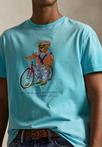 Ανοιχτό μπλε μπλουζάκι με εικόνα αρκούδας με γυαλιά ηλίου και casual ρούχα, που στέκεται δίπλα σε κόκκινο ποδήλατο, φορεμένο με καφέ ζώνη και τζιν.
