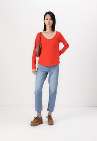 Röd långärmad v-ringad topp, ljusblå raka jeans, bruna platåskor och en brun crossbody-väska mot en vit bakgrund.