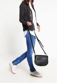 Svart läder crossbody-väska med klaffstängning och metalldetaljer, bärs med en grå jacka, vit skjorta och blå skinny jeans.