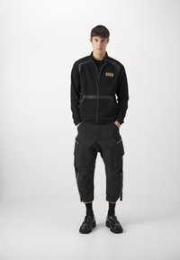 Felpa nera con zip e logo dorato, abbinata a pantaloni cargo neri tapered con dettagli zippati e accenti argentati. Scarpe nere completano l'outfit.