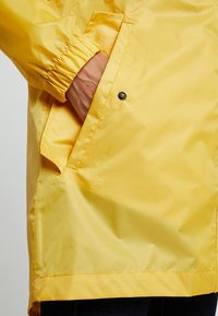 Heldergele regenjas met elastische manchetten, een zijzak met drukknoopsluiting en een gladde, waterdichte stof.