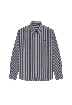 Kvinde står vendt fremad iført rød kasket, stribet knappet skjorte over hvid t-shirt, grå shorts, hvide sokker og grå sneakers mod en ensfarvet baggrund.