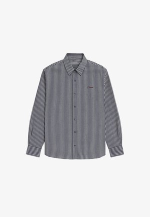 Frau steht nach vorne gerichtet, trägt eine rote Kappe, ein gestreiftes Hemd mit Knöpfen über einem weißen T-Shirt, graue Shorts, weiße Socken und graue Sneakers vor einfachem Hintergrund.