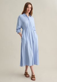 Abito camicia a strisce blu chiaro con chiusura a bottoni, vita elastica, maniche lunghe arrotolate e tasche laterali. Modello abbinato a sandali marroni.