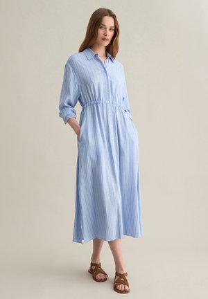 Lichtblauwe gestreepte jurk met knoopsluiting, elastische taille, lange opgekrulde mouwen en zijzakken. Model draagt er bruine sandalen bij.