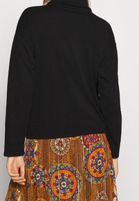 Mujer vistiendo una blusa negra de manga larga y una falda colorida con un patrón de diseños circulares, vista desde atrás.