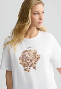 T-shirt bianco in cotone con design di rosa a stampa leopardata e dettagli metallici; logo "LIU JO" sopra la grafica. Maniche corte, vestibilità comoda.
