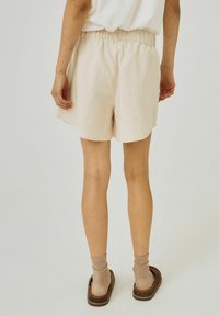 Light beige cotton shorts with an elastic waistband, relaxed fit, and slightly curved hem. Pairing with brown slip-on shoes and socks.