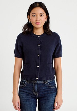 Mujer con un cárdigan azul marino de manga corta y botones, con dos bolsillos delanteros y vaqueros azul oscuro, de pie frente a un fondo liso.